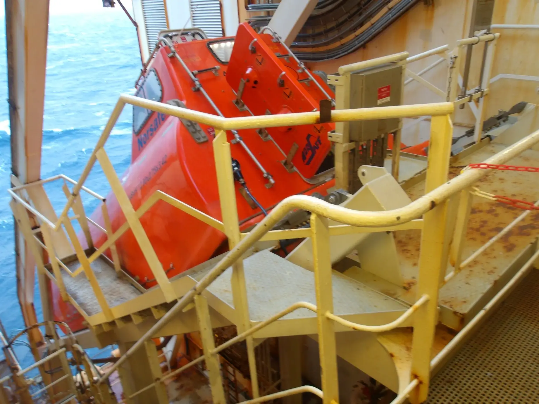 An orange lifeboat is being lowered from a ships deck into the ocean, surrounded by safety equipment and metal structures.