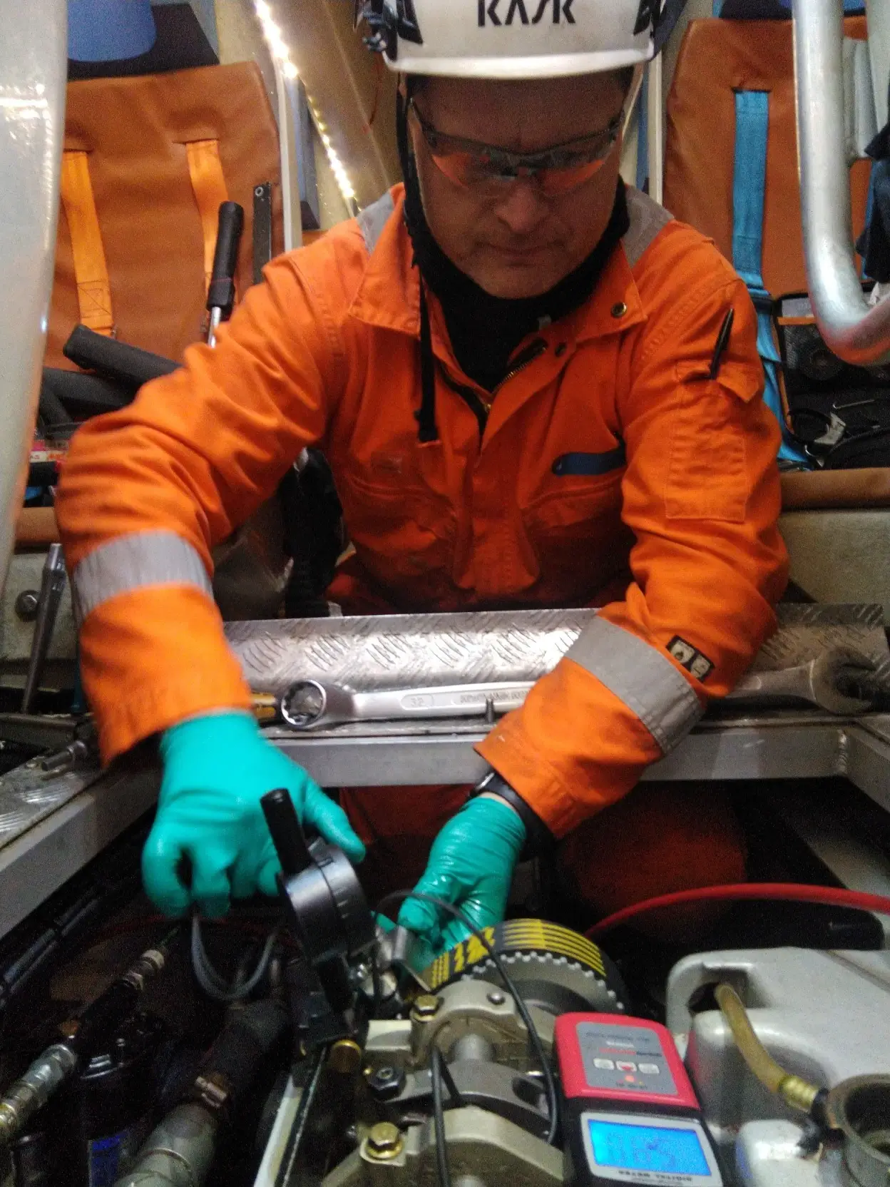 En tekniker fra Agder Motor Service, iført en oransj overdel og grønne hansker, arbeider med maskineri ved hjelp av verktøy inne i et mekanisk rom.