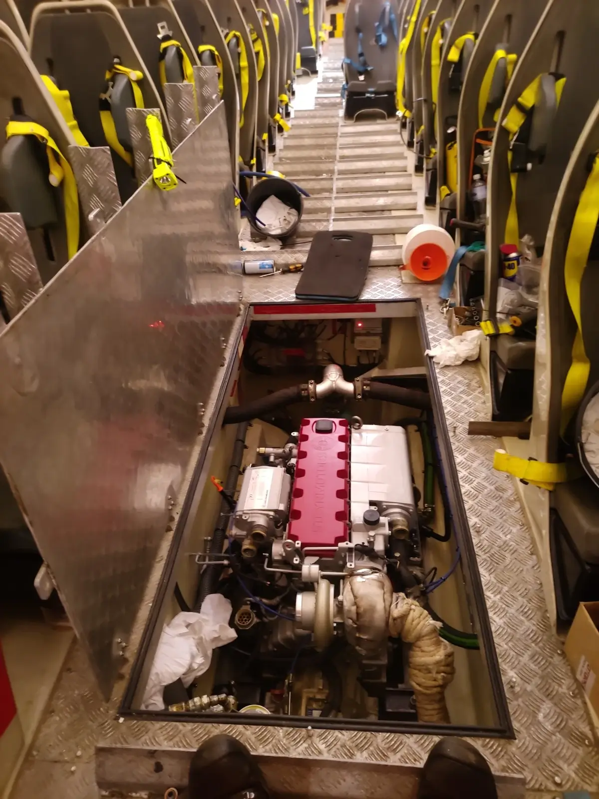 Et livbåtinteriør med seter, der en motorblokk er synlig i et rom på gulvet, omgitt av verktøy og vedlikeholdsutstyr.