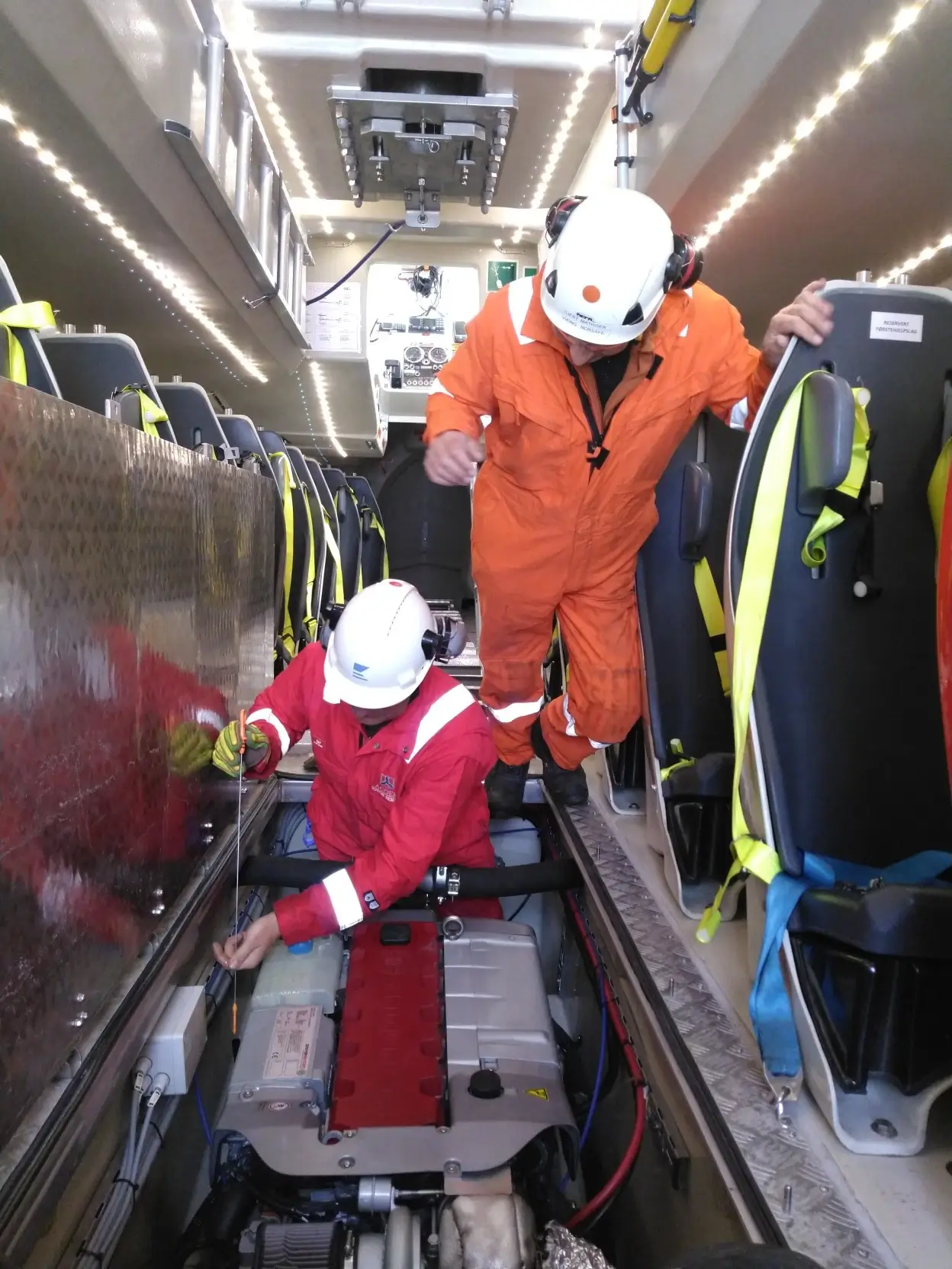 To mekanikere fra Agder Motor Service, utstyrt med hjelmer og verneklær, jobber inne i en bil. Den ene kontrollerer motordelen mens den andre hjelper til ovenfra